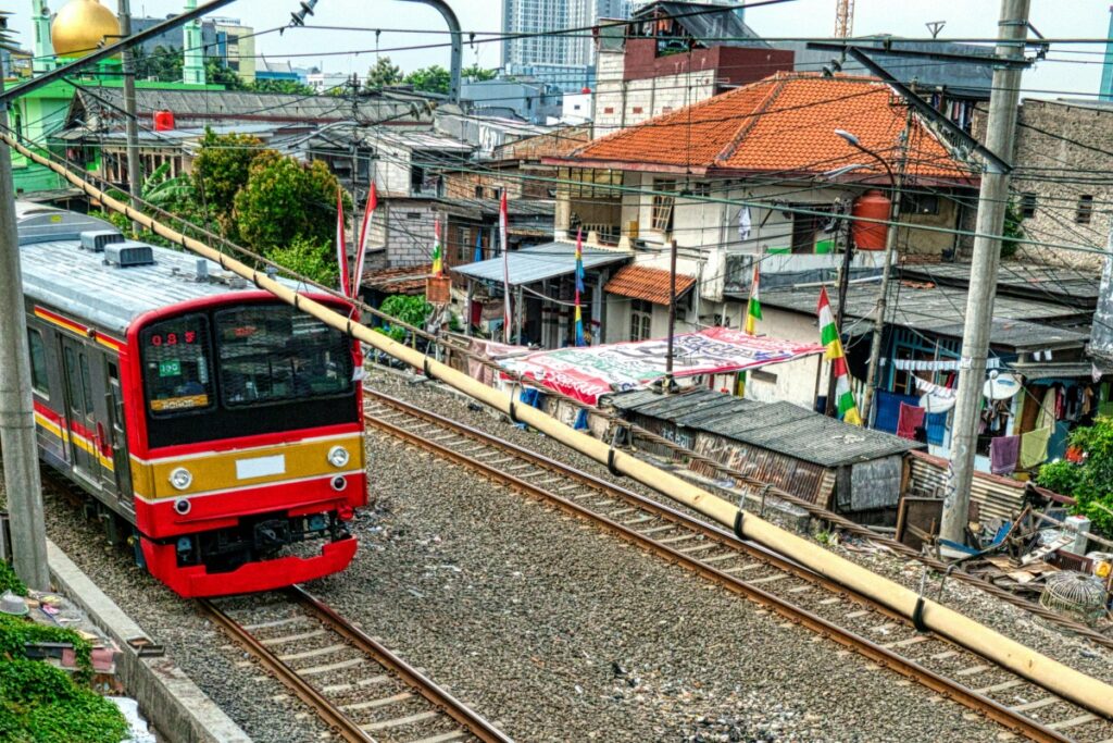 Gambar ini menampilkan sebuah kereta api dengan warna merah, kuning, dan hitam yang sedang melaju di atas rel kereta. Di sekelilingnya, terdapat perkampungan dengan rumah - rumah sederhana, atap - atap yang berwarna beda - beda, serta beberapa bendera yang bergoyang. Di latar belakang, ada gedung - gedung modern yang menonjol.