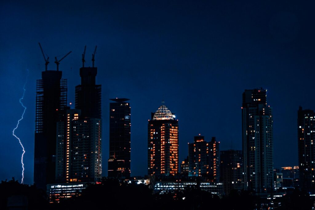 Gambar ini memperlihatkan skyline kota pada malam hari, dengan gedung - gedung tinggi yang bercahaya dan beberapa kran bangunan yang terlihat menanjak ke langit. Tiba - tiba, petir terbit, menambah keindahan dan kehebohan gambar.